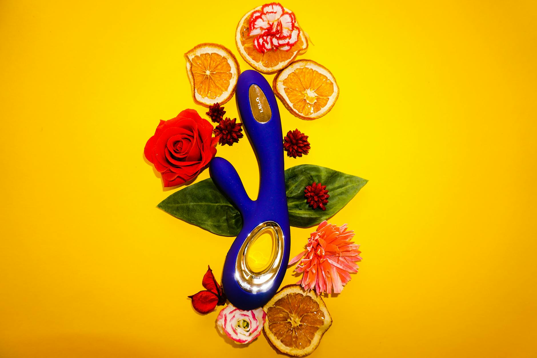 Blue vibrator surrounded by flowers and citrus on yellow background