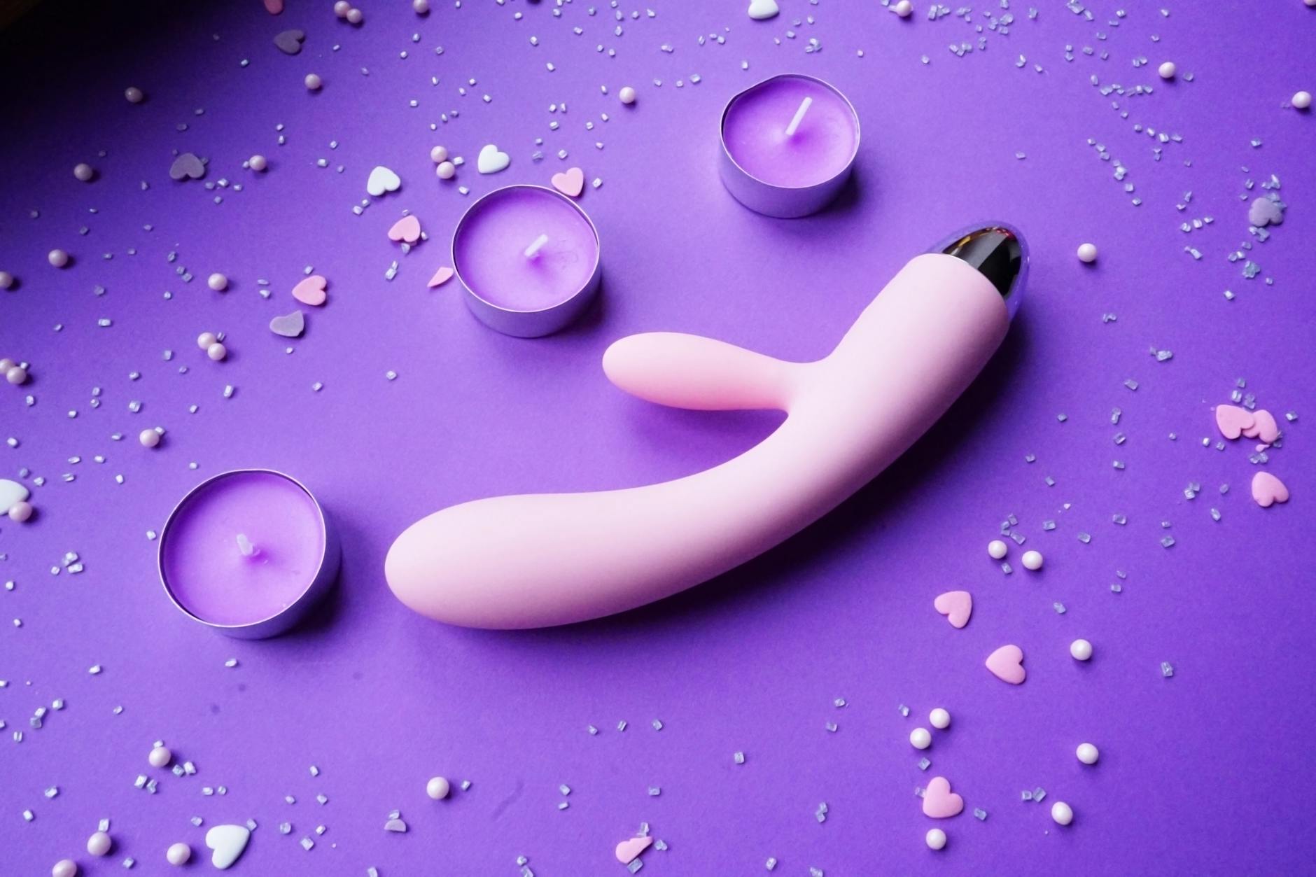 Pink clitoral vibrator surrounded by candles and heart confetti on a romantic purple background